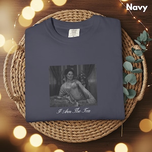 May include: A navy blue crewneck t-shirt featuring a black and white graphic print of a person in a vintage-style dress sitting on a bed. Below the image, the text reads "I Am The Tea" in a white cursive font. The shirt has a Comfort Colors label.