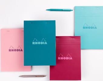 May include: Four rectangular Rhodia notepads in light pink, teal, magenta, and light blue, each featuring the Rhodia logo and two staples at the top. A teal pen is positioned above the notepads, and a silver pen is placed below them.