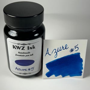 May include: A black bottle of KWZ Ink handmade fountain pen ink with a white label reading KWZ Ink handmade fountain pen ink Azure #5. Next to the bottle is a white card with the handwritten text Azure #5 and a blue ink swatch.