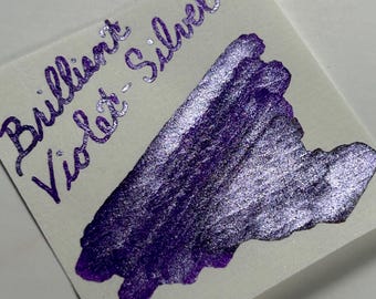 Può includere: Un cartoncino quadrato bianco con una scritta a mano in inchiostro viola metallizzato che recita Brilliant Violet-Silver, accompagnata da un ampio campione di inchiostro viola e argento scintillante e testurizzato.