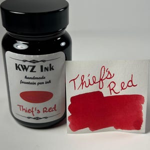 May include: A glass bottle of KWZ Ink handmade fountain pen ink with a black cap and a white label reading "KWZ Ink handmade fountain pen ink" and "Thief's Red". Next to it is a white paper swatch with "Thief's Red" written in red ink.