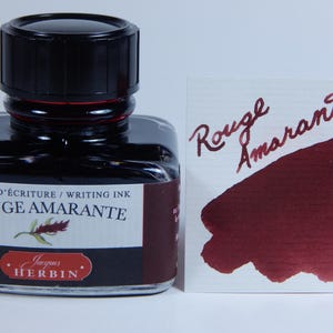 May include: A square glass bottle of Rouge Amarante writing ink with a black cap. The label reads "Rouge Amarante" and "Jacques Herbin." A swatch of the ink displays a deep red color.