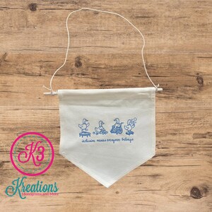 May include: White canvas banner with a blue duck design and the text "inclusion means everyone belongs." The banner is suspended by a white string and wooden dowel. The banner has a triangular shape.