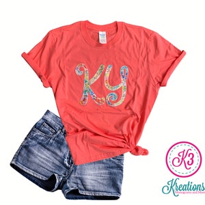 Puede incluir: Una camiseta de color coral con un monograma floral de las letras "KY" en la parte delantera. La camiseta se combina con pantalones cortos de mezclilla.