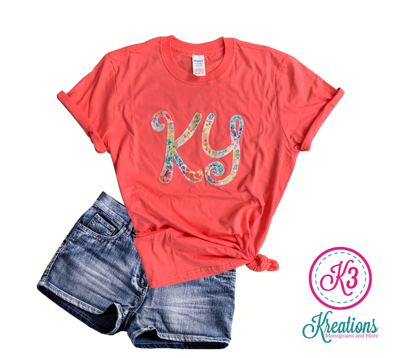 Puede incluir: Una camiseta de color coral con un monograma floral de las letras "KY" en la parte delantera. La camiseta se combina con pantalones cortos de mezclilla.