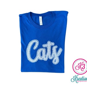 May include: Royal blue t-shirt with a white sparkly "Caty" design. The shirt is folded and the brand tag is visible.