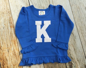 May include: A royal blue long-sleeved shirt with a white ruffle trim around the bottom and sleeves. The shirt features a large white letter "K" in a block font.
