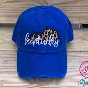 May include: A royal blue distressed baseball cap with a white "kentucky" embroidered on the front. The state of Kentucky is outlined in black and filled with a leopard print pattern.