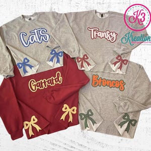 May include: Four grey sweatshirts with different colored text and bows on the sleeves. The text on the sweatshirts reads "Cats", "Transy", "Gaitard", and "Broncos".