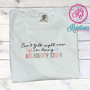 May include: White t-shirt with the text "Can't talk right now I'm doing AUDIOLOGY STUFF" in rainbow colors with a star on each side of the text.