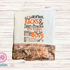 May include: White quilt with a brown and orange camouflage border. The quilt has an embroidered design that says "Rifles, Racks & Deer Tracks are what little boys are made of".