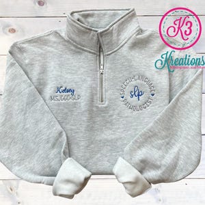 May include: Gray sweatshirt with a zipper and embroidered text that reads "Kelsey MS, CCC-SLP" and a circle with the text "Speech-Language Pathologist" and the abbreviation "SLP" inside the circle.