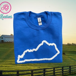 May include: A royal blue sweatshirt with a white outline of the state of Kentucky. The sweatshirt is folded and photographed in front of a green grassy field with a white barn in the background.