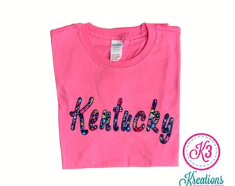 May include: Pink t-shirt with a floral patterned "Kentucky" design in a cursive font. The shirt is folded and the brand tag is visible.