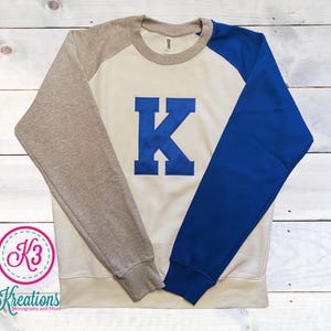 May include: A raglan-sleeve sweatshirt with a cream-colored body, a gray left sleeve, and a blue right sleeve. A large blue letter "K" is centered on the front. The sweatshirt is on a white wooden surface. The logo "K3 Kreations Monograms and More" is in the lower left corner.