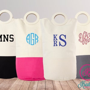 May include: Four white canvas laundry baskets with wooden handles. Each basket has a different embroidered design: MNS, AGR, KS, and DFF. The baskets are half black, half pink, half gray, and half white.