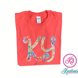 Puede incluir: Una camiseta de color rosa coral con un monograma bordado de colores "KY" en la parte delantera. El monograma est&aacute; hecho de una variedad de telas y colores, incluyendo rosa, azul, verde, amarillo y blanco. La camiseta est&aacute; doblada y colocada sobre un fondo blanco.