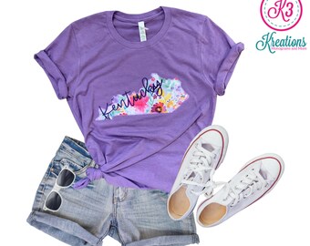 May include: Purple t-shirt with a floral design of the state of Kentucky and the word "Kentucky" in cursive. The shirt is paired with denim shorts and white sneakers.
