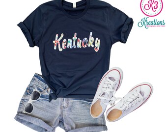 May include: A navy blue t-shirt with the word "Kentucky" in colorful patterned letters. The shirt is paired with denim shorts and white sneakers.