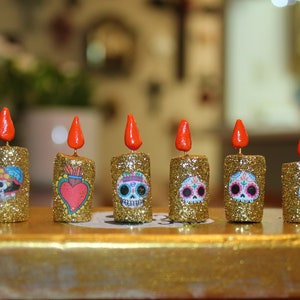 May include: Six gold glitter candles with red wicks and colorful Day of the Dead skull designs. The candles are arranged in a row on a gold surface.