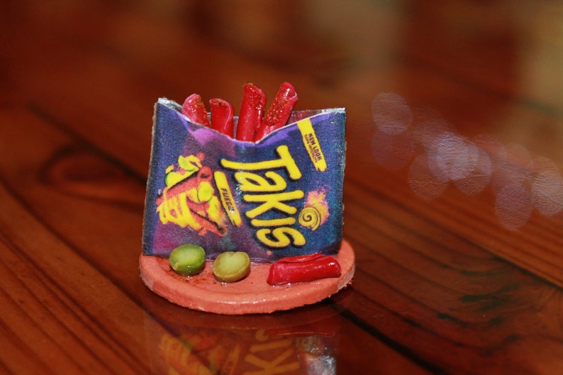 May include: A miniature replica of a Takis Fuego snack bag with red and yellow flames on the front. The bag is filled with red and orange chips and is sitting on a red clay base with two green olives.