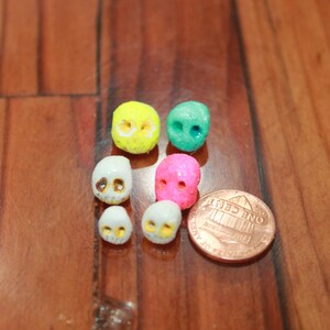 May include: A collection of small, colorful skull-shaped beads in yellow, green, pink, and white. A penny is included for size reference. The beads are arranged on a wooden surface.