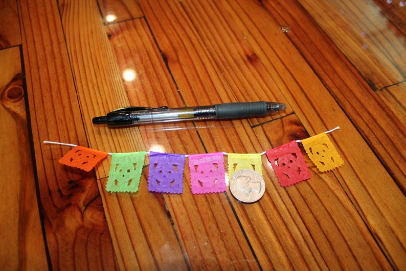 May include: A colorful paper banner with cut-out designs hangs on a wooden surface. The banner is made of tissue paper and features a variety of colors, including orange, green, purple, pink, and yellow. A silver coin is visible in the center of the banner.