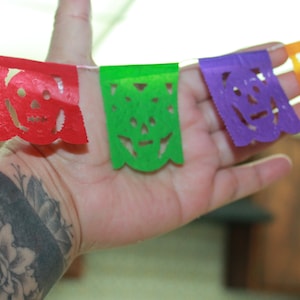 May include: A colorful paper banner with cut-out skull designs in red, green, purple, and yellow. The banner is hanging from a string and is being held by a hand with a black and grey floral tattoo.