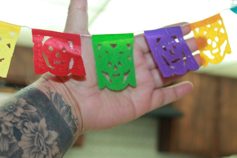 May include: A colorful paper banner with cut-out skull designs in red, green, purple, and yellow. The banner is hanging from a string and is being held by a hand with a black and grey floral tattoo.