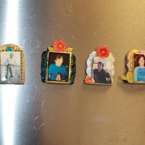 May include: Four framed photographs with gold trim and colorful accents. Each frame features a different person. The frames are attached to a silver surface.