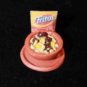 May include: A miniature clay bowl filled with a mixture of yellow and brown food, resembling chili and cheese. A small bag of Fritos chips sits behind the bowl.