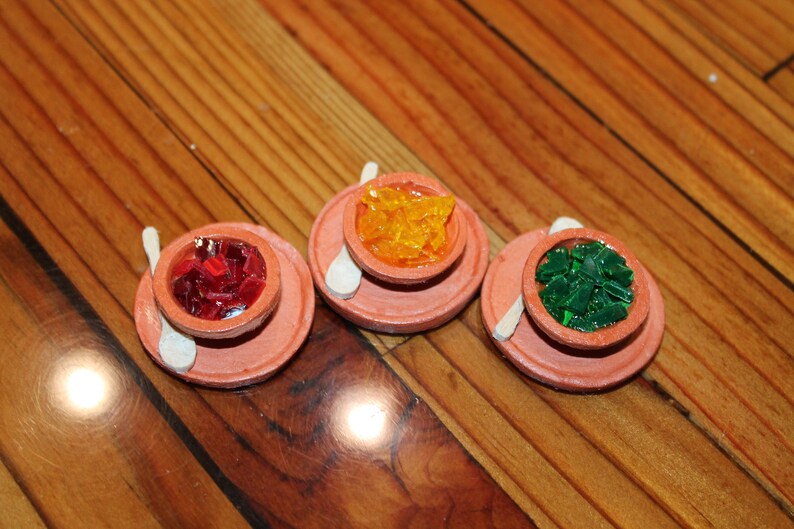 May include: Three small terracotta bowls with spoons, each filled with colorful gelatin cubes. The bowls are arranged in a row on a wooden surface. The gelatin cubes are red, yellow, and green.