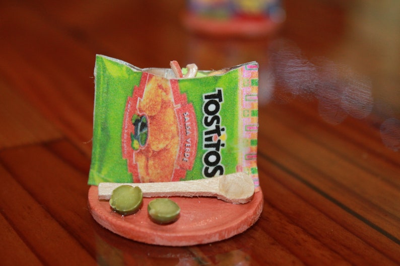 May include: A miniature scene of a small wooden spoon resting on a red clay dish with two green olives. The dish is decorated with a small bag of Tostitos tortilla chips with the text "Tostitos Salsa Verde" printed on the bag.