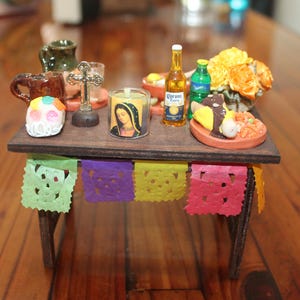 May include: Miniature Day of the Dead altar with a sugar skull, candle, cross, food, and drinks. The altar is decorated with colorful papel picado banners and sits on a wooden table.