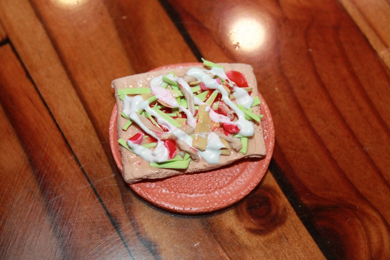 May include: A miniature clay pizza with green, red, and white toppings on a small orange plate.