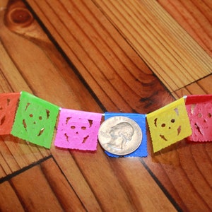 May include: A colorful paper banner with five flags featuring a skull design. The flags are orange, green, pink, yellow, and red. A quarter is included for scale.