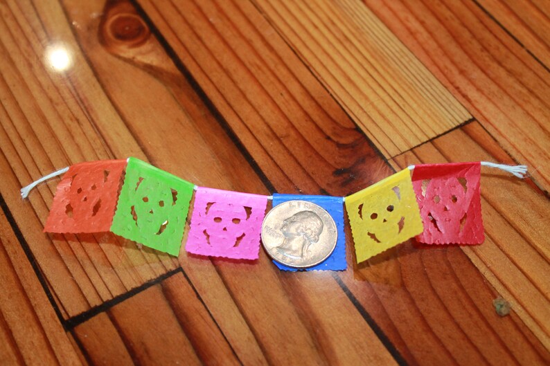 May include: A colorful paper banner with five flags featuring a skull design. The flags are orange, green, pink, yellow, and red. A quarter is included for scale.