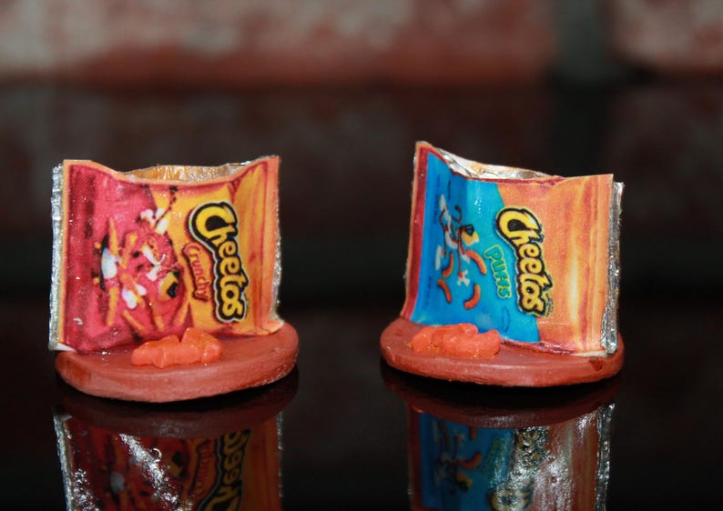 May include: Two miniature Cheeto-themed ring holders. One is red and orange with the text "Cheetos Crunchy" and the other is blue and orange with the text "Cheetos Puffs".