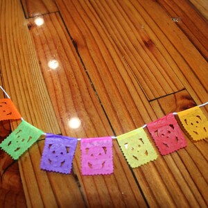 May include: A colorful paper banner with cut-out skull designs hangs on a wooden surface. The banner is made of tissue paper and is strung together with white thread. The colors of the banner are orange, green, purple, pink, yellow, and red.