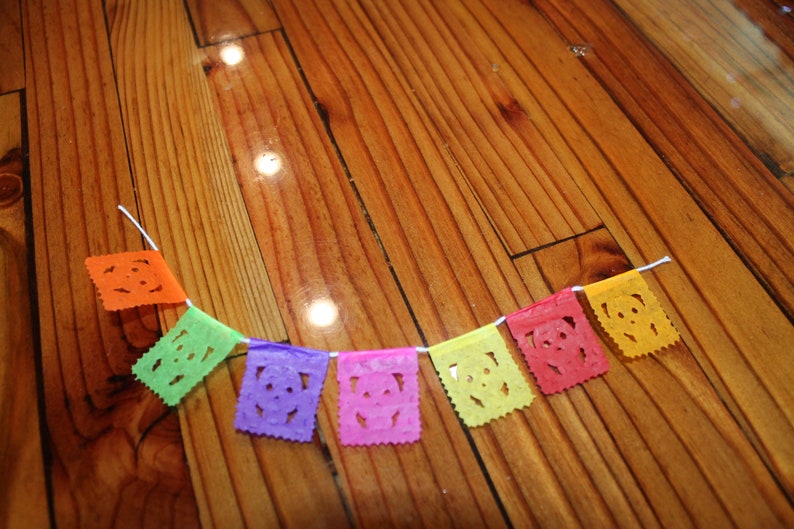 May include: A colorful paper banner with cut-out skull designs hangs on a wooden surface. The banner is made of tissue paper and is strung together with white thread. The colors of the banner are orange, green, purple, pink, yellow, and red.
