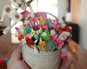 May include: A decorative basket filled with colorful paper flowers, held in a hand. A skeleton figure with butterfly wings is perched above the basket. The basket is made of woven material.