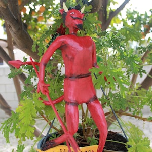 May include: A red devil figurine with horns, a mustache, and a trident. The figurine is standing on a wooden base with the words "EL DIABLITO" on a yellow banner. The figurine is in a blue pot with green leaves.