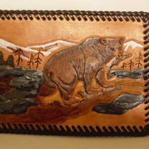 May include: A brown leather wallet with a tooled design of a bear in a mountain landscape. The wallet has a brown stitched border.