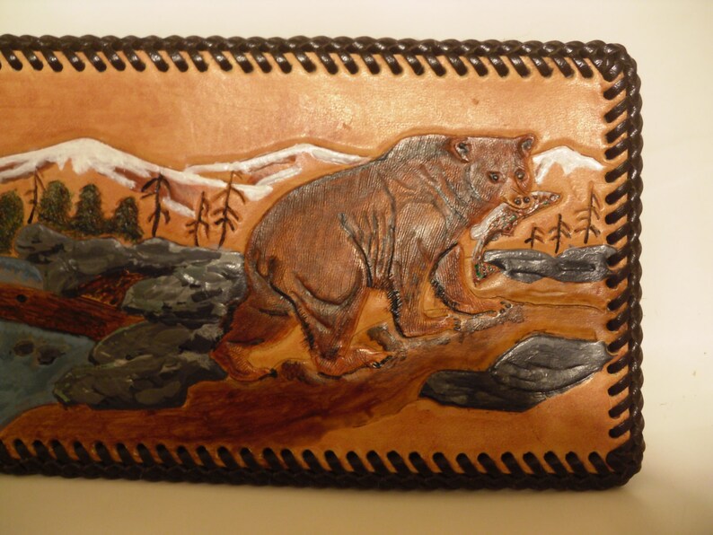 May include: A brown leather wallet with a tooled design of a bear in a mountain landscape. The wallet has a brown stitched border.
