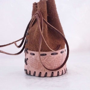 May include: A brown leather pouch with a drawstring closure and a decorative pine cone design. The pouch is made of suede and has a brown leather strap.