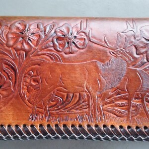 Peut inclure: Un portefeuille en cuir marron avec un motif ciselé de deux élans et de motifs floraux. Le portefeuille a une bordure en cuir tressé.