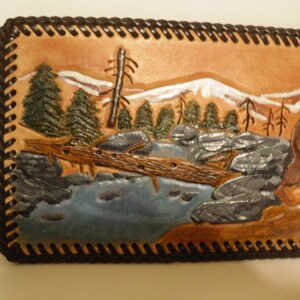May include: A leather wallet with a carved scene of a deer standing in a river with a fallen log and trees in the background. The wallet has a brown leather border with a decorative stitching pattern.