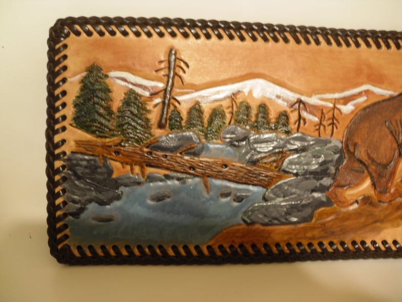 May include: A leather wallet with a carved scene of a deer standing in a river with a fallen log and trees in the background. The wallet has a brown leather border with a decorative stitching pattern.