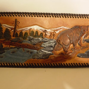 May include: A brown leather wall hanging with a carved image of a bear walking through a mountain scene. The bear is brown and the mountains are white with a blue river in the foreground.