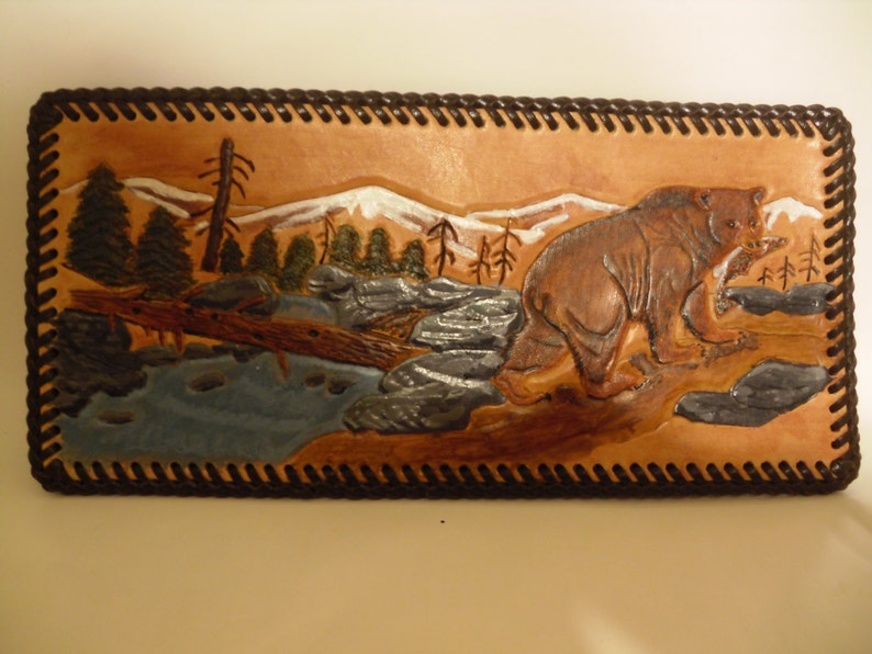 May include: A brown leather wall hanging with a carved image of a bear walking through a mountain scene. The bear is brown and the mountains are white with a blue river in the foreground.